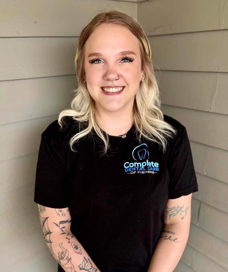 The image features a woman standing against a wall, smiling at the camera, wearing a black t-shirt with a graphic on the front and visible tattoos on her arms.